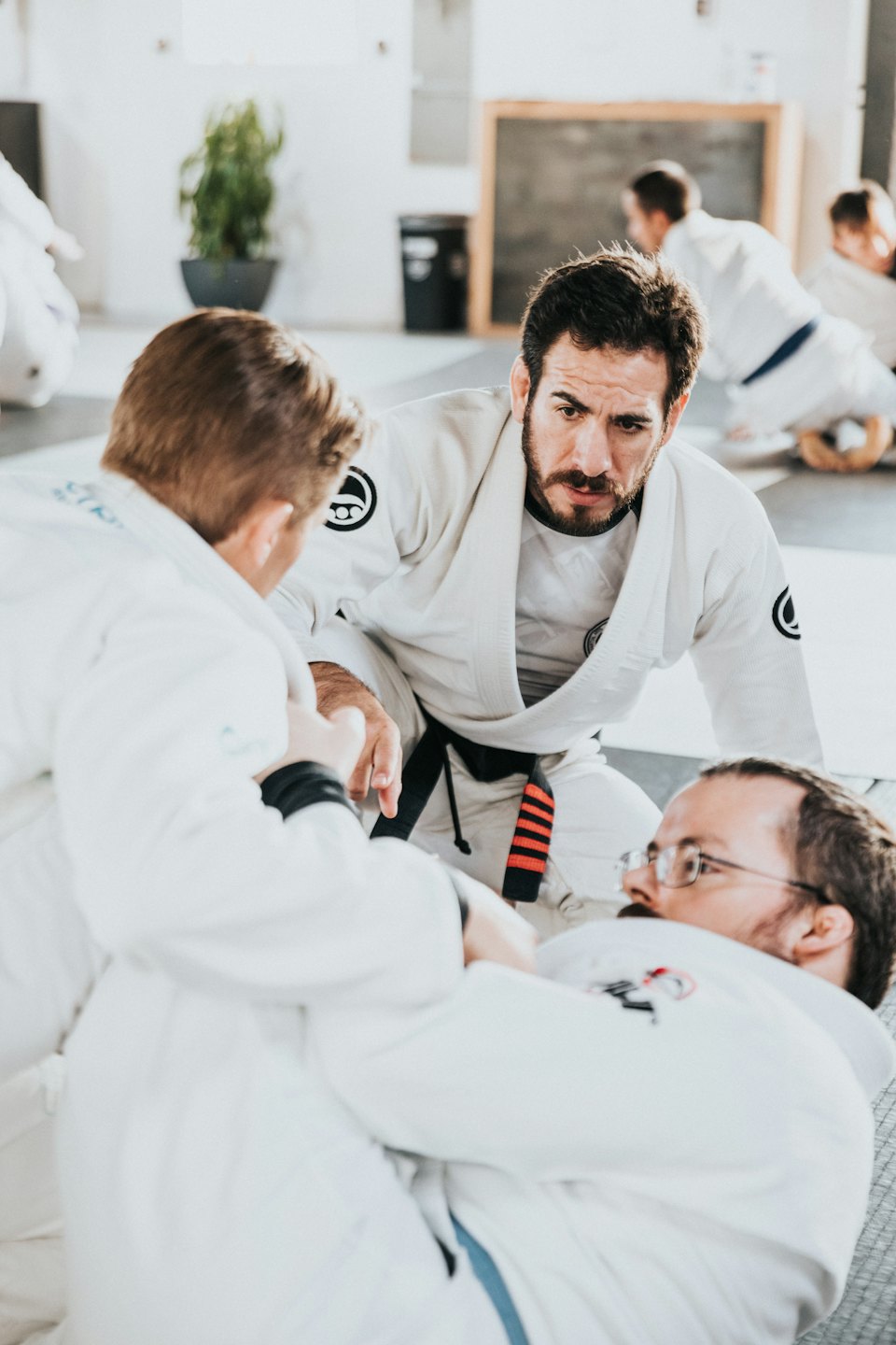 Bearded instructor watches students practice Brazilian Jiu-Jitsu on mats.