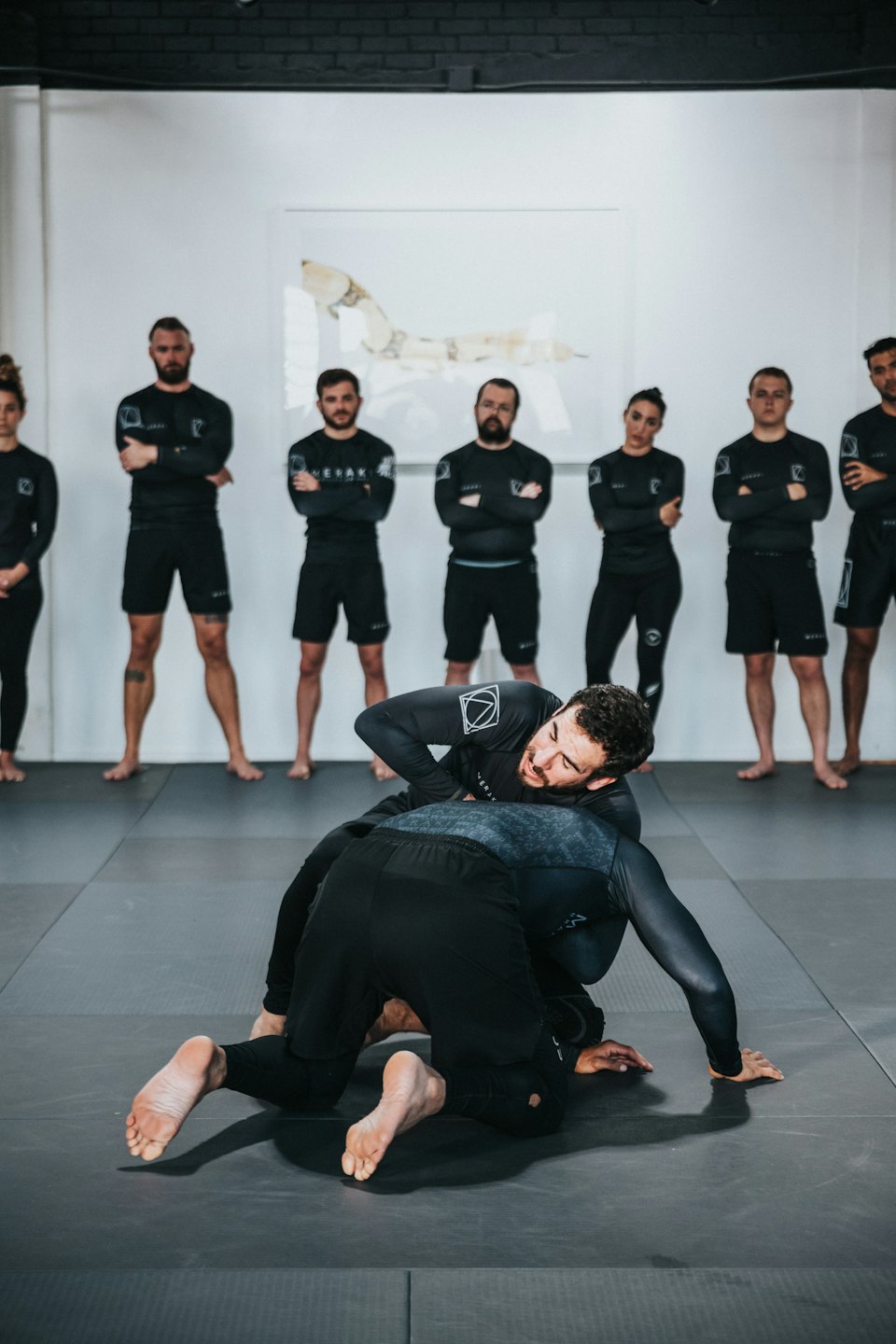 Athletes grappling on gym mat with observers in background.